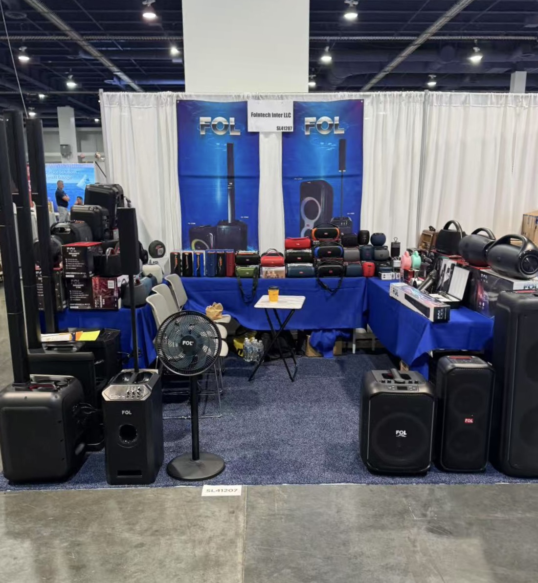 Stand de FOL Audio en la feria ASD 2025, exhibiendo una amplia gama de equipos de audio: altavoces portátiles, columnas acústicas y sistemas de sonido, dispuestos en mesas cubiertas de mantel azul con pancartas de la marca.